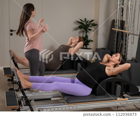 Two pregnant women are doing Pilates on a reformer. The instructor teaches prenatal yoga. Two pregnant women are doing Pilates on a reformer. The instructor teaches prenatal yoga. 112036873