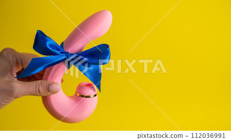 Woman holding curved pink vibrator with blue ribbon on yellow background. Copy space. Woman holding curved pink vibrator with blue ribbon on yellow background. Copy space. 112036991