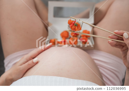 A pregnant woman sits on the sofa and eats rolls from a box. Food delivery. Top view of the tummy. A pregnant woman sits on the sofa and eats rolls from a box. Food delivery. Top view of the tummy. 112036995