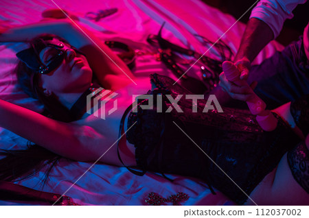 A woman in a leather mask and handcuffs lies on a bed in a blue-red light. Man holding dildo.  112037002