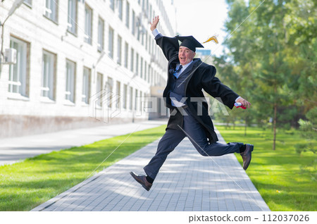 Old happy man in graduation gown jumping outdoors and holding diploma. 112037026