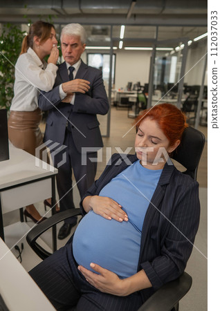 Elderly Caucasian man and woman gossiping behind their sleeping pregnant colleague in the office. Vertical photo.  112037033