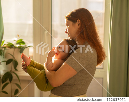 Caucasian woman tenderly holds her newborn son while standing near the window. Caucasian woman tenderly holds her newborn son while standing near the window. 112037051