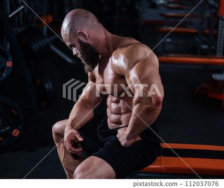 A muscular bald man in shorts is resting on a bench after a workout. Bodybuilder showing off his shape in the gym. A muscular bald man in shorts is resting on a bench after a workout. Bodybuilder showing off his shape in the gym. 112037076