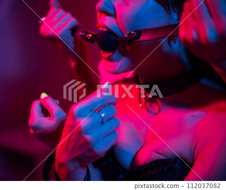 A man gags a woman in a pink blue neon light in the bedroom. BDSM concept. A man gags a woman in a pink blue neon light in the bedroom. BDSM concept. 112037082