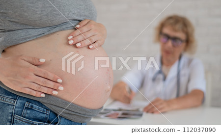 Doctor obstetrician gynecologist at his desk in the background. Close-up of a pregnant woman's belly. Doctor obstetrician gynecologist at his desk in the background. Close-up of a pregnant woman's belly. 112037090