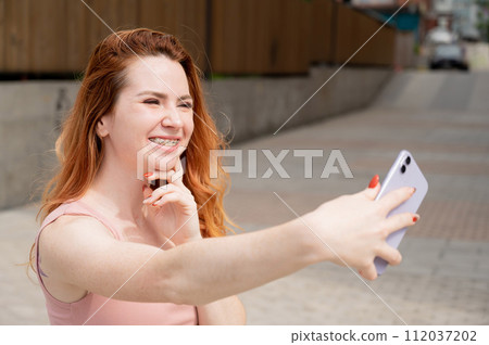 Young woman with braces on her teeth smiles and takes a selfie on a smartphone outdoors. Young woman with braces on her teeth smiles and takes a selfie on a smartphone outdoors. 112037202