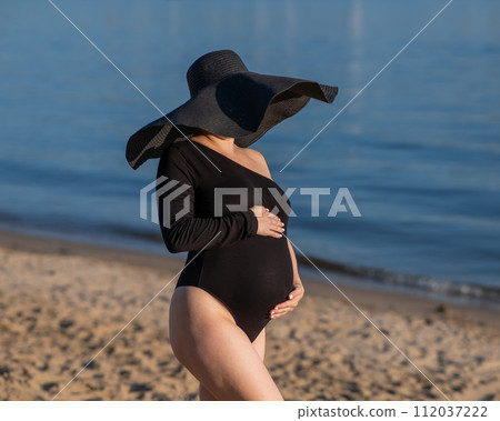 Pregnant woman in a large straw hat and black swimsuit posing on the beach. Pregnant woman in a large straw hat and black swimsuit posing on the beach. 112037222