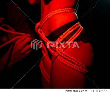 Man tightens ropes using Japanese shibari technique on woman's body in red light.  112037355