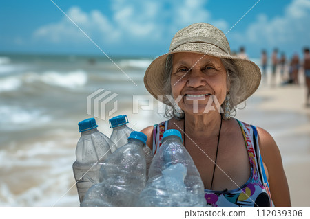 Smiling elderly woman holding crumpled plastic bottles on the beach. 112039306