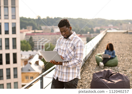 Business man dressed standing on roof top outdoors and typing on modern laptop. 112040257
