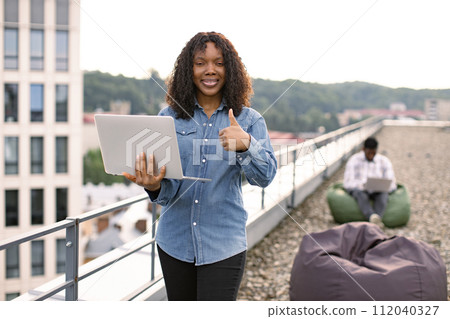 Female using laptop outdoors, male colleague works sitting in bag chair. 112040327