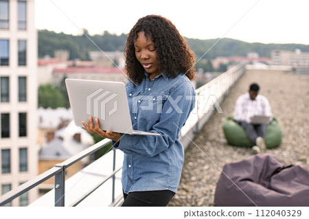Female using laptop outdoors, male colleague works sitting in bag chair. 112040329