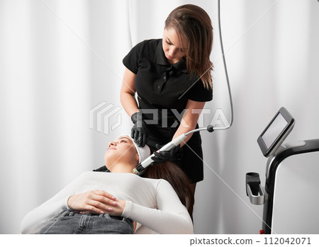 Doctor cosmetologist using radiofrequency microneedling device while performing rejuvenation treatment in cosmetology clinic. Woman lying near modern equipment and having skincare procedure. Doctor cosmetologist using radiofrequency microneedling device while performing rejuvenation treatment in cosmetology clinic. Woman lying near modern equipment and having skincare procedure. 112042071