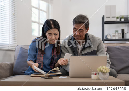 Asian senior couple checking and calculate financial bill together on sofa involved in financial paperwork, paying tax online using laptop at living room home Asian senior couple checking and calculate financial bill together on sofa involved in financial paperwork, paying tax online using laptop at living room home 112042638