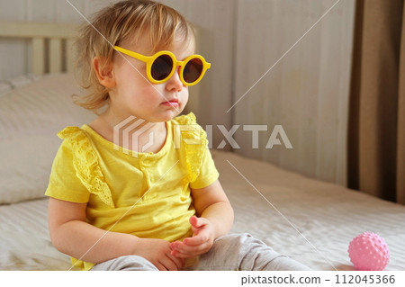 Child playing with colorful plastic toys on beige cozy bed in sunny white bedroom. Educational game for baby and toddler. Young girl play indoors. Wearing sunglasses. Kid preparing to vacation, travel Child playing with colorful plastic toys on beige cozy bed in sunny white bedroom. Educational game for baby and toddler. Young girl play indoors. Wearing sunglasses. Kid preparing to vacation, travel 112045366