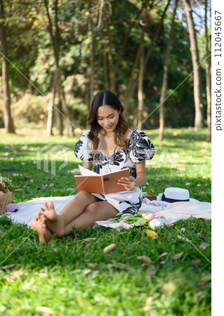 Full length of happy young woman sitting on grass with barefoot and reading book 112045667