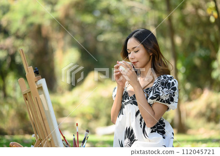 Peaceful young woman sitting in front of easel in the garden and drinking hot coffee Peaceful young woman sitting in front of easel in the garden and drinking hot coffee 112045721