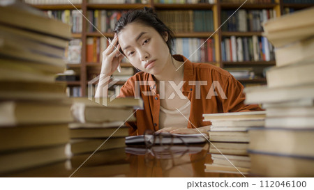 Portrait of exhausted asian woman student sitting behind stacks of books in a library Portrait of exhausted asian woman student sitting behind stacks of books in a library 112046100