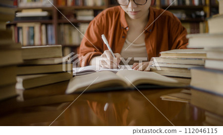 Female student in eyeglasses taking notes, studying books in college library 112046115