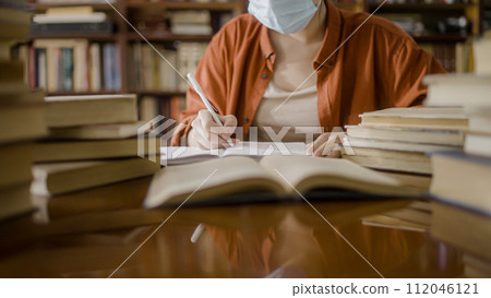 Asian female writer in face mask conducting research at library, making notes Asian female writer in face mask conducting research at library, making notes 112046121