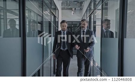 Male business partners walking in office corridor, discussing future cooperation Male business partners walking in office corridor, discussing future cooperation 112046808