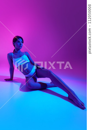 Young slim woman in white lingerie posing sitting on floor in blue neon light against gradient purple background. Fitness and sport Young slim woman in white lingerie posing sitting on floor in blue neon light against gradient purple background. Fitness and sport 112050008
