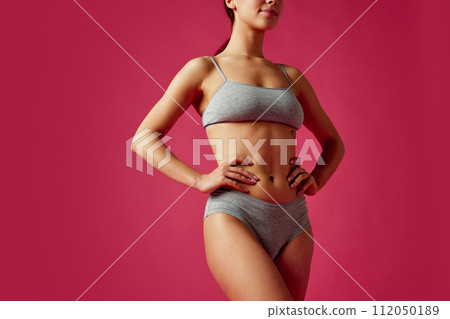 Cropped portrait of young woman in grey lingerie posing holds hands on hips against vivid pink studio background. 112050189