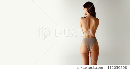 Rear view of half-naked woman in beige underwear standing hug herself against grey studio background. Body positivity Rear view of half-naked woman in beige underwear standing hug herself against grey studio background. Body positivity 112050208