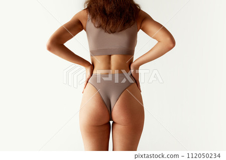 Rear view. Cropped portrait of young woman in lingerie holds hands on hips. Model with slim figure against grey studio background. 112050234