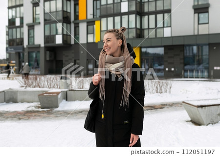 A young pretty woman with blond dreadlocks dressed warmly walks along the street in winter 112051197
