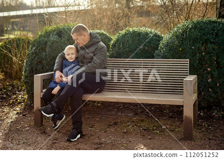 Autumnal Family Affection: Father, 40 Years Old, and Son - Beautiful 8-Year-Old Boy, Seated in the Park. Autumnal Family Affection: Father, 40 Years Old, and Son - Beautiful 8-Year-Old Boy, Seated in the Park. 112051252