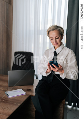 Vertical shot of smiling middle-aged businesswoman using smartphone sitting on office desk, touching screen, typing message on mobile phone, browsing Internet, social media, shopping online. 112052986