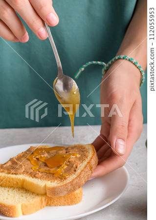 Woman's hands puting honey with spoon on a toast with peanut butter to make a sandwich for breakfast 112055953