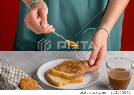 Woman's hands puting honey with spoon on a toast with peanut butter to make a sandwich for breakfast 112055971