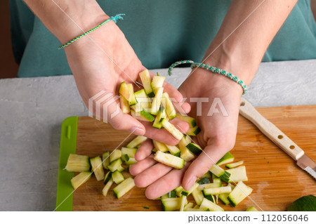 Authentic female hands holding cutting zucchini by cubes on wooden cutting board on kitchen table Authentic female hands holding cutting zucchini by cubes on wooden cutting board on kitchen table 112056046