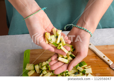 Authentic female hands holding cutting zucchini by cubes on wooden cutting board on kitchen table Authentic female hands holding cutting zucchini by cubes on wooden cutting board on kitchen table 112056047
