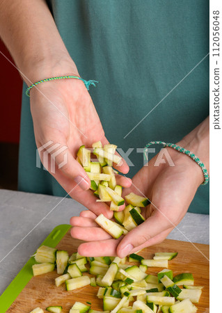Authentic female hands holding cutting zucchini by cubes on wooden cutting board on kitchen table Authentic female hands holding cutting zucchini by cubes on wooden cutting board on kitchen table 112056048
