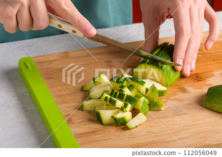 Authentic female hands cutting zucchini by cubes on wooden cutting board on kitchen table 112056049