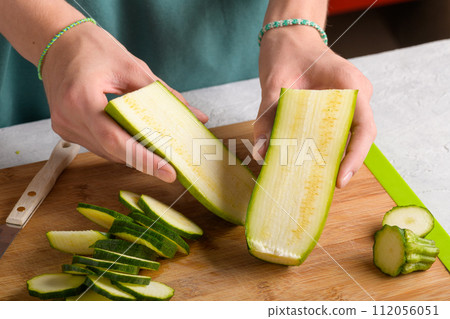 Woman's hands cutting zucchini on wooden cutting board at home, preparing healthy food, close up Woman's hands cutting zucchini on wooden cutting board at home, preparing healthy food, close up 112056051
