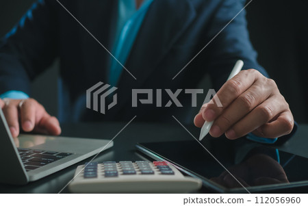 Businessman in black suit working on laptop computer and tablet and calculator, Hand Hands touch on tablet at office with dark background, Online working, Close up, Copy space. Businessman in black suit working on laptop computer and tablet and calculator, Hand Hands touch on tablet at office with dark background, Online working, Close up, Copy space. 112056960
