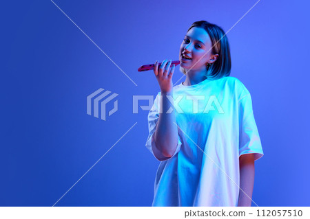 Gel portrait of young girl in casual clothes recording voice message mobile phone against blue studio background in neon light Gel portrait of young girl in casual clothes recording voice message mobile phone against blue studio background in neon light 112057510
