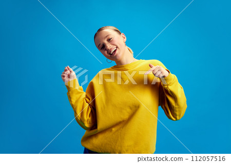 Positive, beautiful, smiling young girl in yellow sweatshirt dancing, having fun against blue studio background 112057516