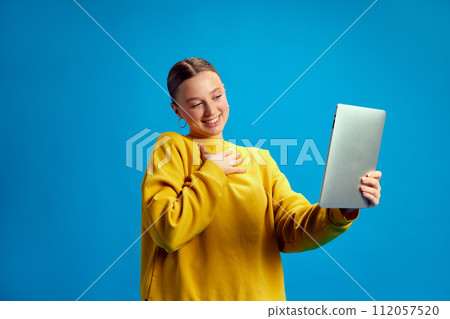 Young girl, student in yellow sweatshirt holding tablet and having online video call, lesson against blue studio background 112057520