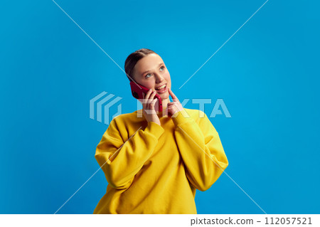 Young beautiful girl with nude makeup, wearing yellow sweatshirt, talking on mobile phone against blue studio background 112057521