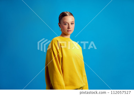 Portrait of beautiful young girl with ponytail, nude makeup, wearing yellow sweatshirt, standing against blue studio background Portrait of beautiful young girl with ponytail, nude makeup, wearing yellow sweatshirt, standing against blue studio background 112057527