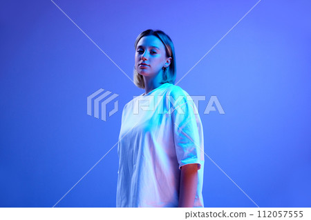 Gel portrait of beautiful young girl in white t-shirt standing, posing against blue studio background in neon light Gel portrait of beautiful young girl in white t-shirt standing, posing against blue studio background in neon light 112057555
