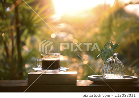 Coffee glass cup on thick old book on wooden table with plant glass vase under sunlight Coffee glass cup on thick old book on wooden table with plant glass vase under sunlight 112058031