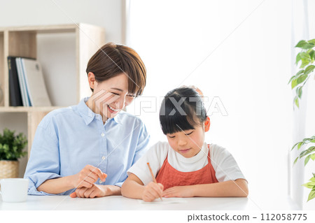 A girl doing her homework and her mother watching over her. 112058775