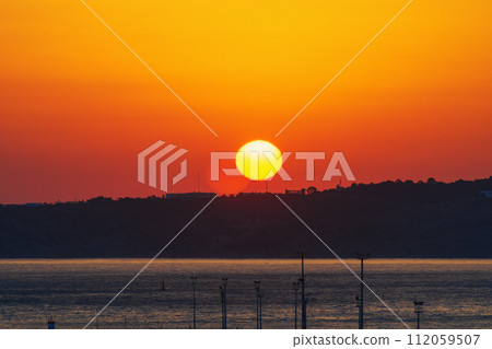 Crimson sunrise over the hill near the seaport. The moment of the beginning of a new day, a symbol of the onset of changes and future achievements. Abstraction, symbolism and realism 112059507